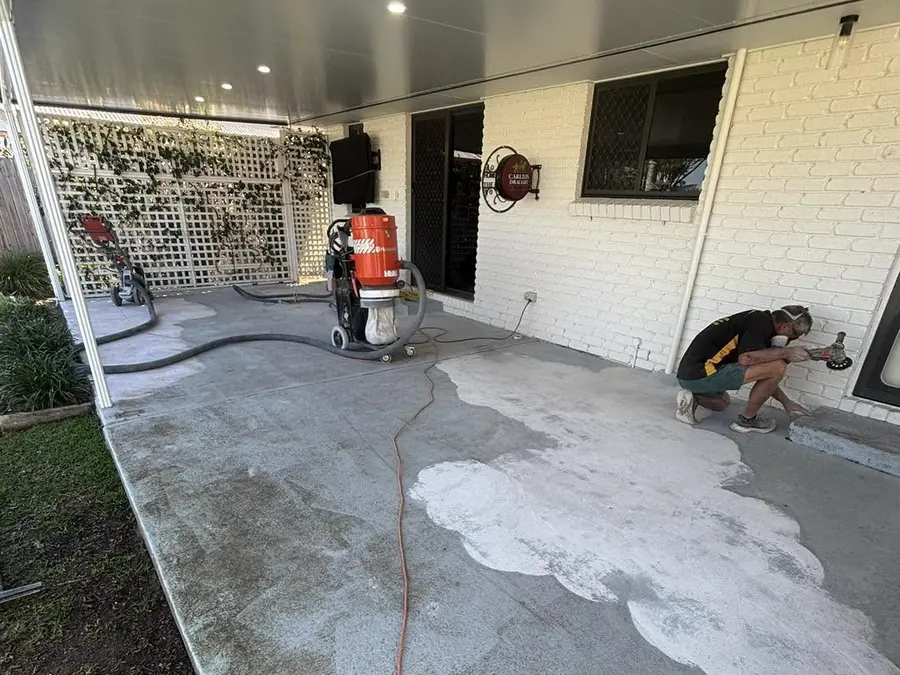 An undercover patio mid-grind: bare concrete, a dust extractor and a worker grinding along the wall