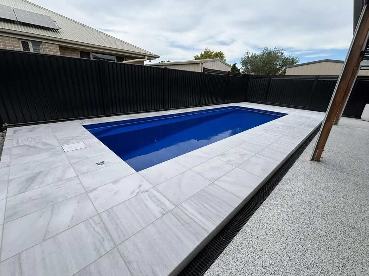 Pale pool surround and tiled coping around a blue plunge pool, beside a black Colorbond fence